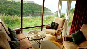 A room with large windows offering a view of a green landscape and a body of water. There are two patterned armchairs and a small round table with a woven base.