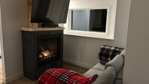 A living room with a fireplace, a television on a wooden stand, a window, and a grey sofa with a plaid throw blanket. - Ski, Bike and family fun at "The Future" cottage.  (Jay)