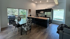 A modern kitchen with a central island, black cabinets, and a stainless steel refrigerator. There is a dining area with a table and chairs, and large windows offering a view of the outdoors. - Ski, Bike and family fun at "The Future" cottage.  (Jay)