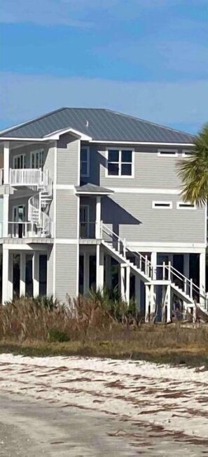 Une maison à deux étages située en bord de mer, dotée d’un balcon et d’un escalier menant à l’entrée principale, avec une plage de sable en premier plan.