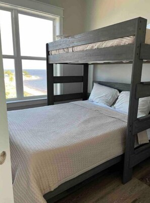 Un lit superposé avec une couvre-lit et des oreillers blancs, placé près d’une fenêtre donnant sur une plage.