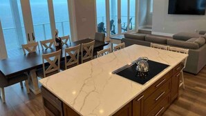 A modern kitchen with a marble countertop, wooden cabinets, and a dining area with chairs. - SandyFeet Beach House Retreat-Gulf Front Home at Shell Point Beach.  (Crawfordville)