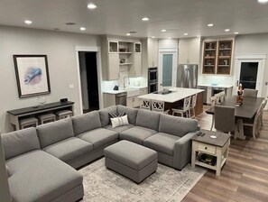 Un salon moderne avec un canapé d’angle gris, un petit pouf et une table basse. On y trouve également un coin repas avec une table et des chaises blanches, ainsi qu’une cuisine avec des armoires blanches et des appareils en acier inoxydable.
