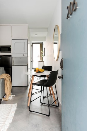 Une cuisine compacte équipée d’un four à micro-ondes, d’un four et d’un réfrigérateur. On y trouve un coin repas avec une table, des chaises et un saladier rempli de fruits. Une porte bleue est visible sur la droite.