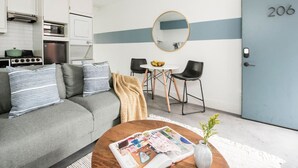 Ett kompakt bostadsutrymme med ett litet kök, en grå soffa med randiga kuddar, en rund spegel, ett matbord med två stolar och en dörr som är märkt med nummer 206.