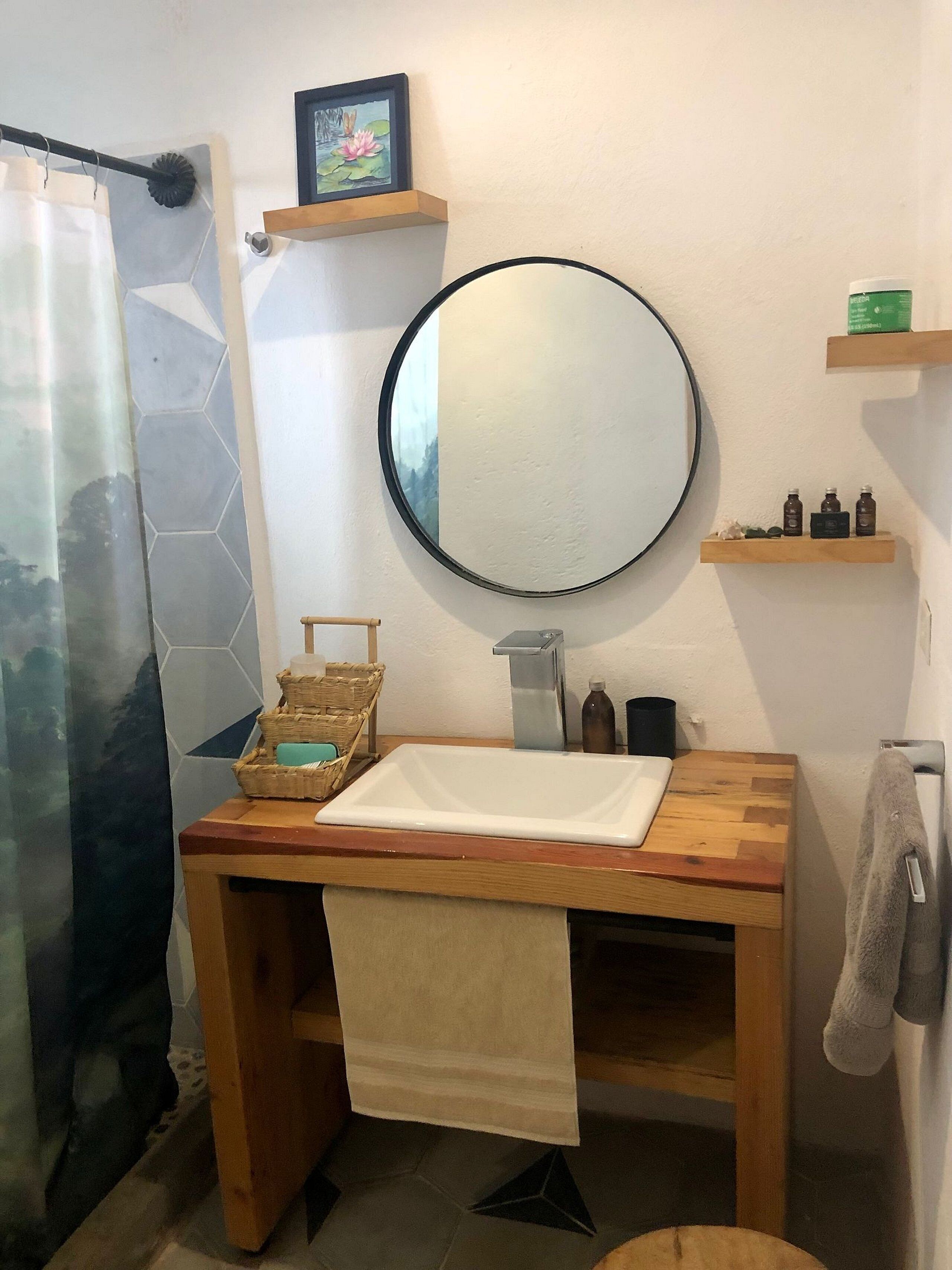 A bathroom with a wooden vanity, a round mirror, a shower curtain, a towel rack, and a small shelf with toiletries.