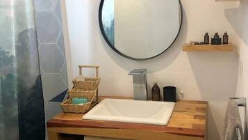 A bathroom with a wooden vanity, a round mirror, a shower curtain, a towel rack, and a small shelf with toiletries.