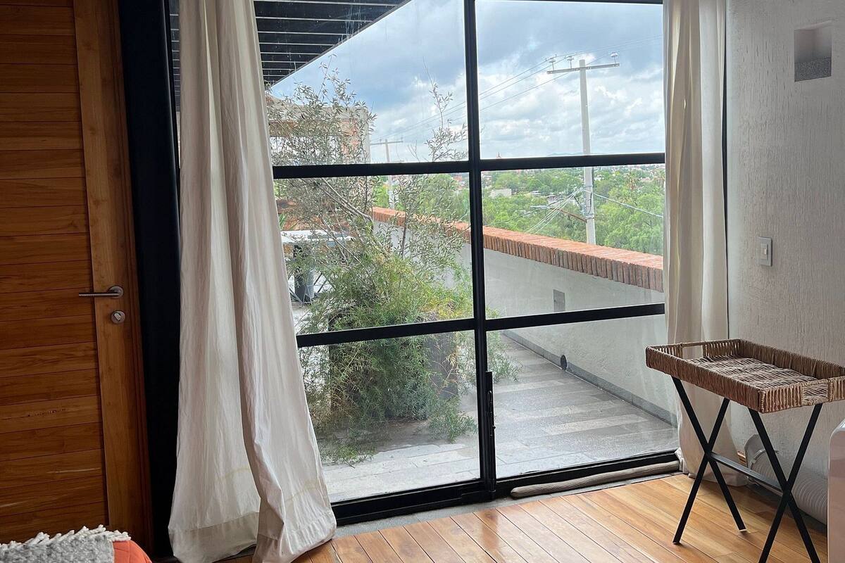 A room with a large sliding glass door, a wooden floor, a bed with a grey and orange blanket, and a woven chair.