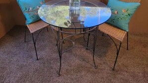 Une table ronde en verre, des chaises en osier, un vase rempli de fleurs et, par la fenêtre, une vue sur les arbres.