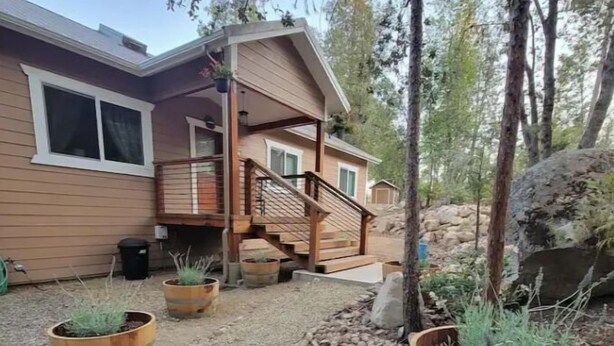 Une maison en bois avec un porche couvert et un escalier menant à une terrasse. Deux grands jardinières en bois, ornées de plantes, se trouvent le long du chemin de gravier.