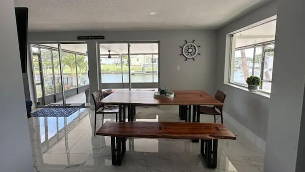 A dining area with a wooden table and chairs, a ship wheel decoration, and large windows offering views of a body of water and greenery.