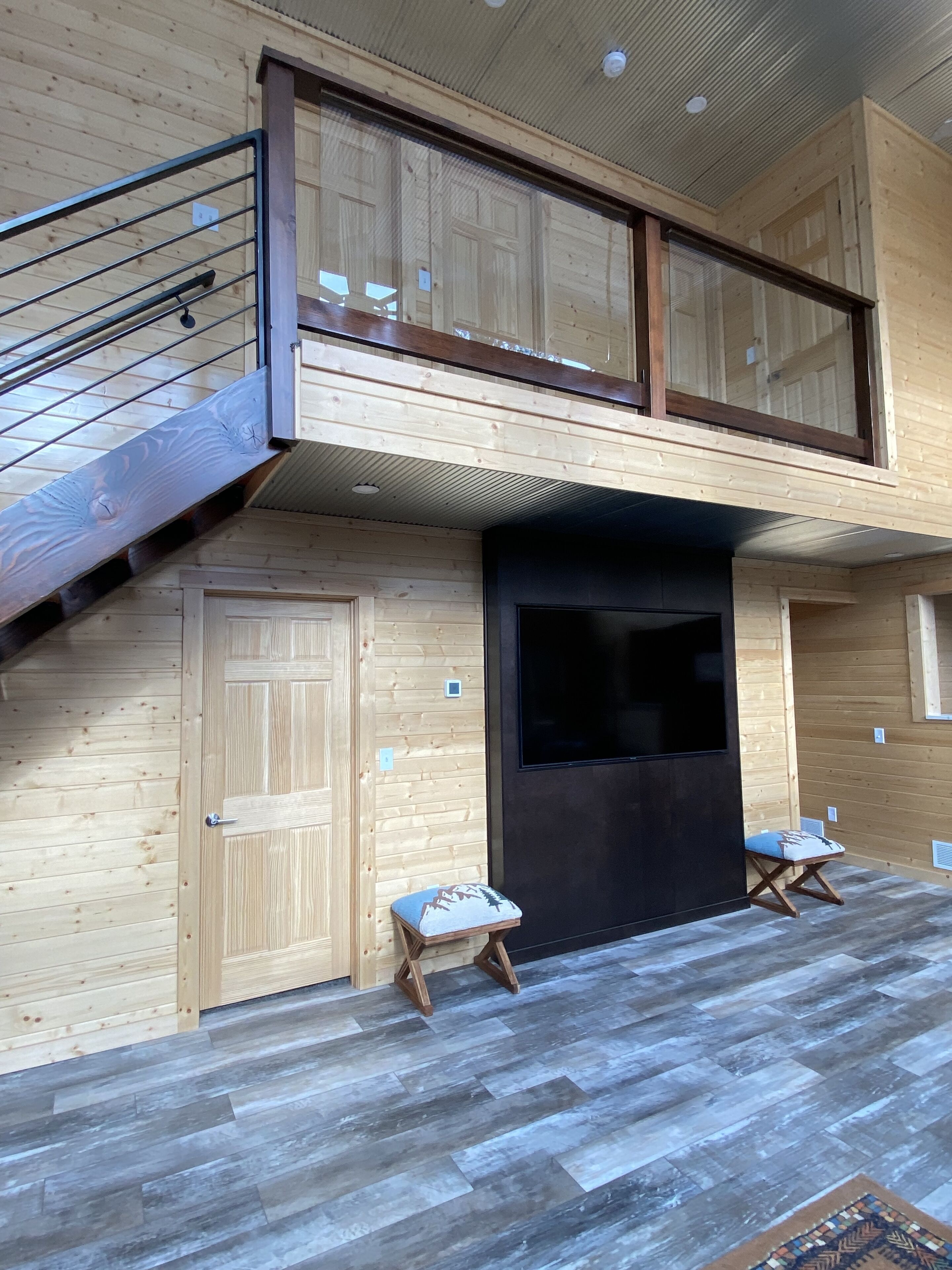 A wooden interior with a staircase leading to a balcony, a large dark-framed window, and two small stools.
