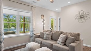 Uma sala de estar com um sofá bege, duas almofadas iguais, uma janela com cortinas, um objeto decorativo na parede e uma porta com luminária.