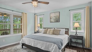 Quarto com uma cama grande, duas mesas de cabeceira, ventilador de teto e janelas com cortinas.