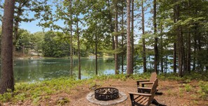 A forested area with a fire pit and a wooden chair near a lake. - Tranquil Waters 30 Night Min on Tibbets Lake (Traverse City)