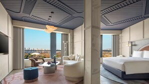 Quarto de hotel moderno com uma cama grande, um sofá, uma mesa de centro e vista para a paisagem urbana.