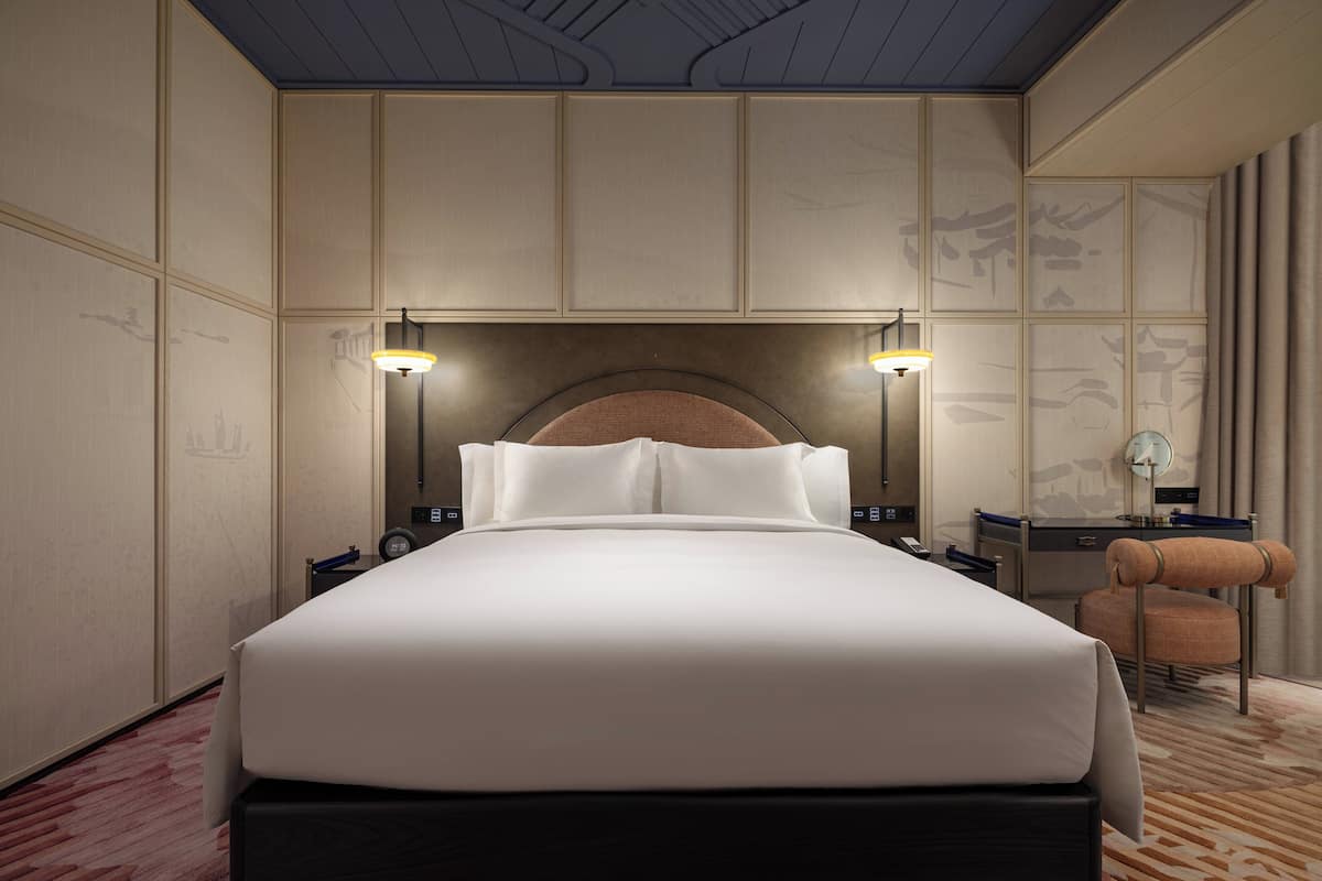Ein Hotelzimmer mit einem großen Bett, zwei Nachttischen, einem Beistelltisch, einem Sessel und einer Decke mit geometrischem Muster.