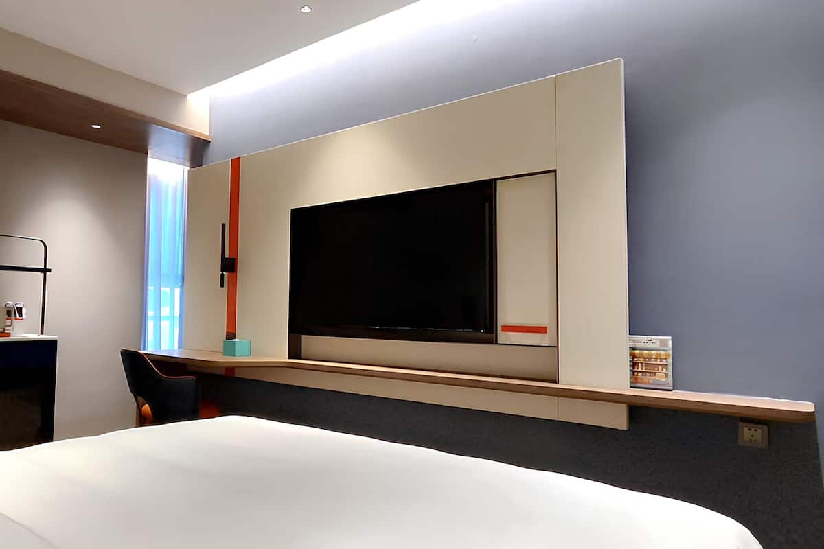A modern hotel room with a large bed, a flat-screen TV mounted on the wall, a desk with a chair, and a small shelf with books.