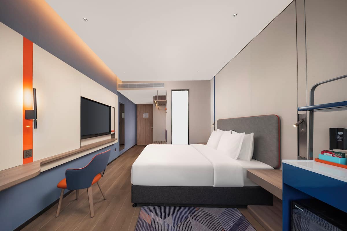 Una habitación de hotel moderna con una cama grande, un televisor montado en la pared, un escritorio con una silla y una pequeña área de cocina.