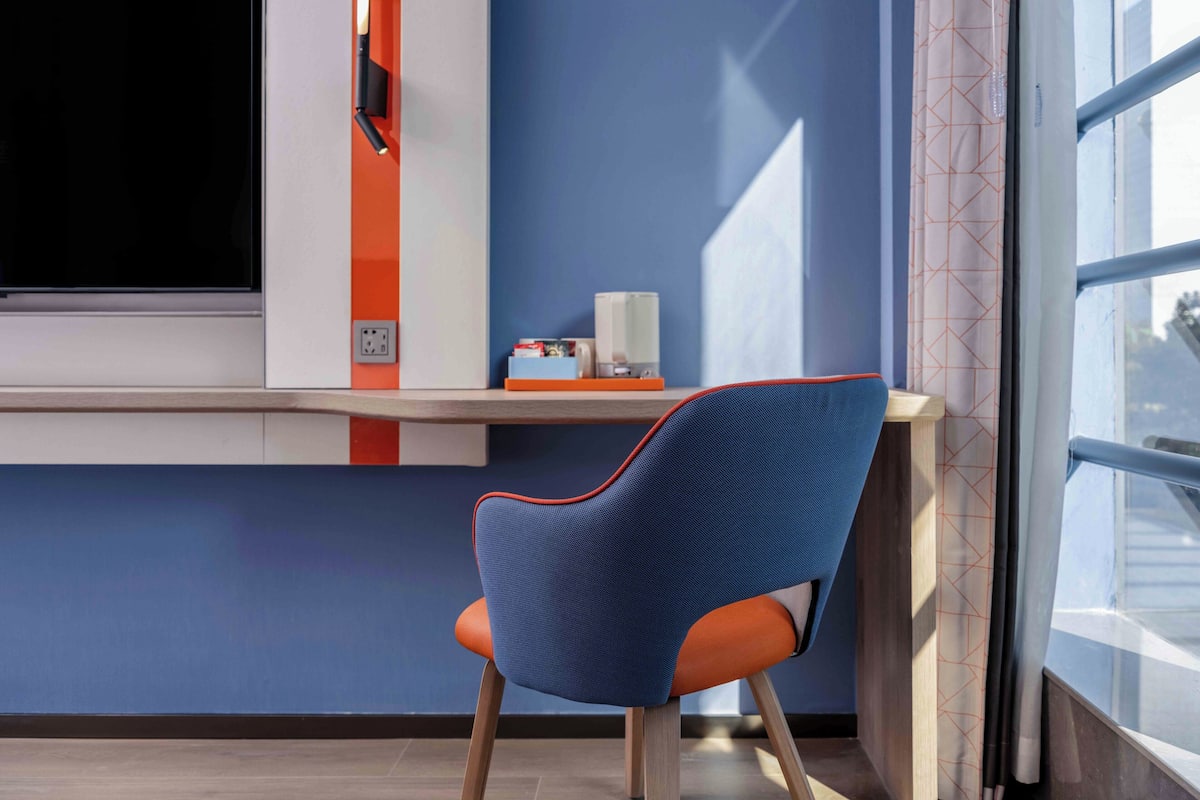A modern room with a blue wall, a window with a view, a chair with orange and blue upholstery, and a shelf with a white object.