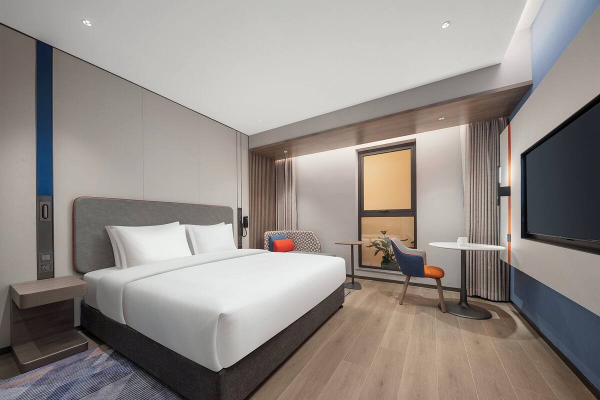 A modern hotel room with a large bed, a desk, a chair, and a TV.