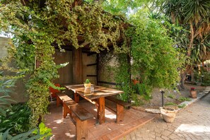 Una mesa de madera al aire libre con bancos, rodeada de abundante follaje y un sendero de ladrillo.
