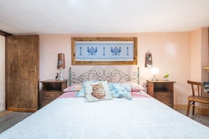 Ein Schlafzimmer mit einem Bett, einer Holztür, einem Nachttisch mit Lampe, einem Stuhl und einem Wandbild.