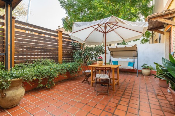 Un patio con suelo de baldosas, una mesa de madera con sillas, un paraguas blanco y plantas en macetas.