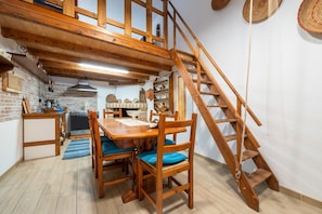 Eine Holztreppe führt zu einer oberen Ebene, einem Essbereich mit Holzsesseln und einem Tisch sowie eine Küche mit Herd und Regalen.