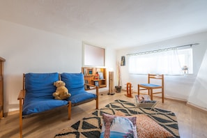 Ein Wohnzimmer mit einem blauen Sofa, einem Holzstuhl, einem Bücherregal und einem Fenster mit Vorhängen.