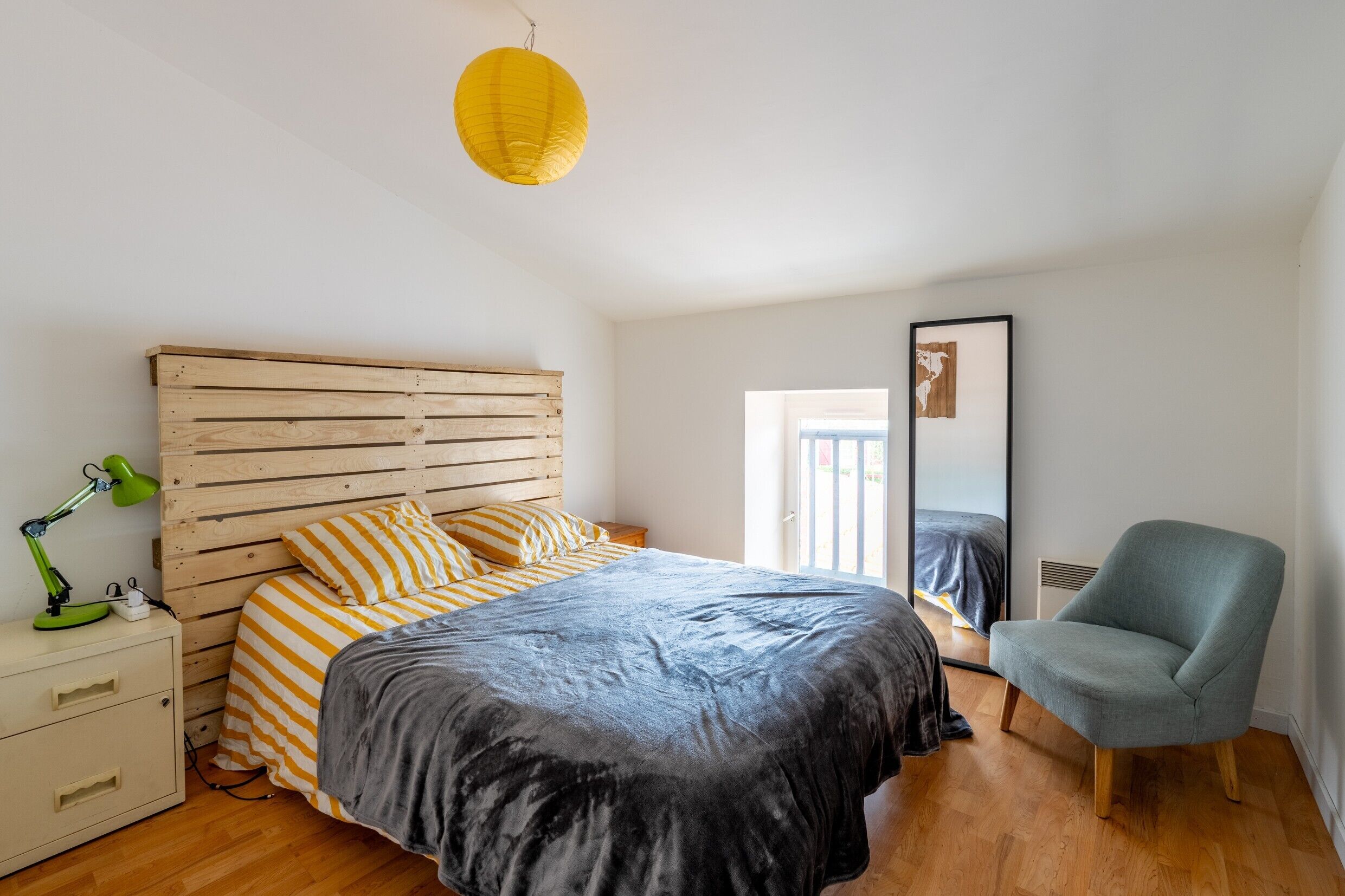 Una camera da letto con testiera in legno, un letto con lenzuola a righe, una sedia verde, un comodino con una lampada e uno specchio a parete.