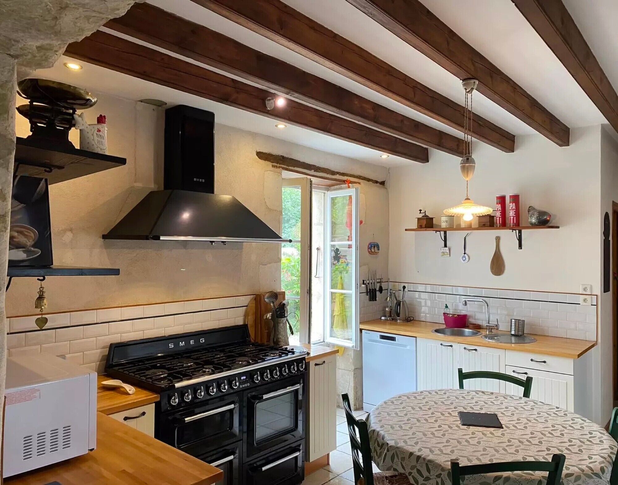 Une cuisine équipée d’une cuisinière, d’un four et de poutres en bois, une table à manger avec des chaises, et une fenêtre offrant une vue agréable.