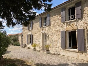 A stone building with multiple windows and shutters, a gravel driveway, and potted plants.