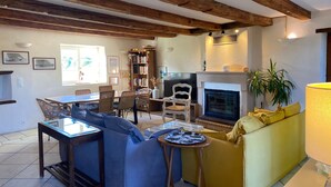 A living room with a fireplace, wooden beams, a sofa, a dining table, and a bookshelf.