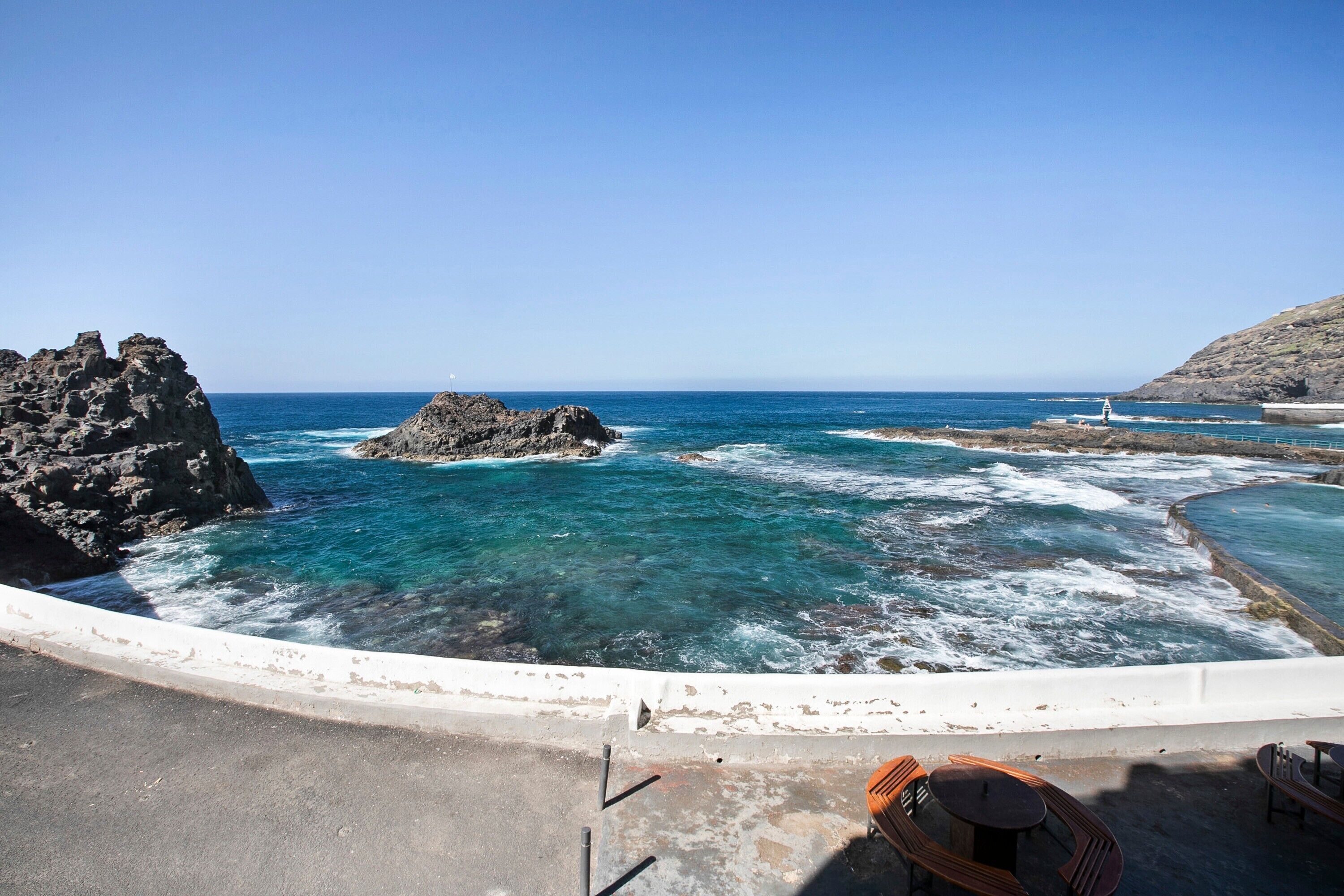 Vista costeira com formações rochosas, água azul límpida e uma estrutura branca curva.