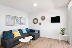 Una sala de estar moderna con un sofá seccional gris, una mesita blanca, un televisor de pantalla plana montado en la pared y una planta en maceta sobre una mesita auxiliar.
