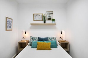 Un dormitorio con una cama, dos mesitas de noche, repisas en la pared y cuadros enmarcados.