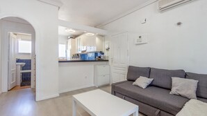 A modern living room with a grey sofa, a white coffee table, and a kitchen area with white cabinets and blue tiles.
