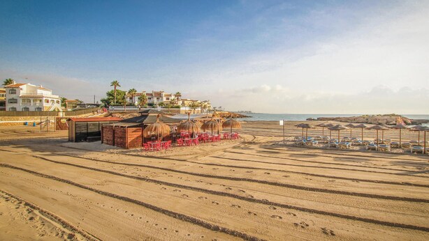 A beach with rows of sun loungers and a small building with red chairs.