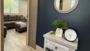 Un couloir avec un mur sombre, un miroir rond, une petite console en bois sur laquelle sont posés une horloge et une plante, et une vue sur un salon avec un canapé et une fenêtre.