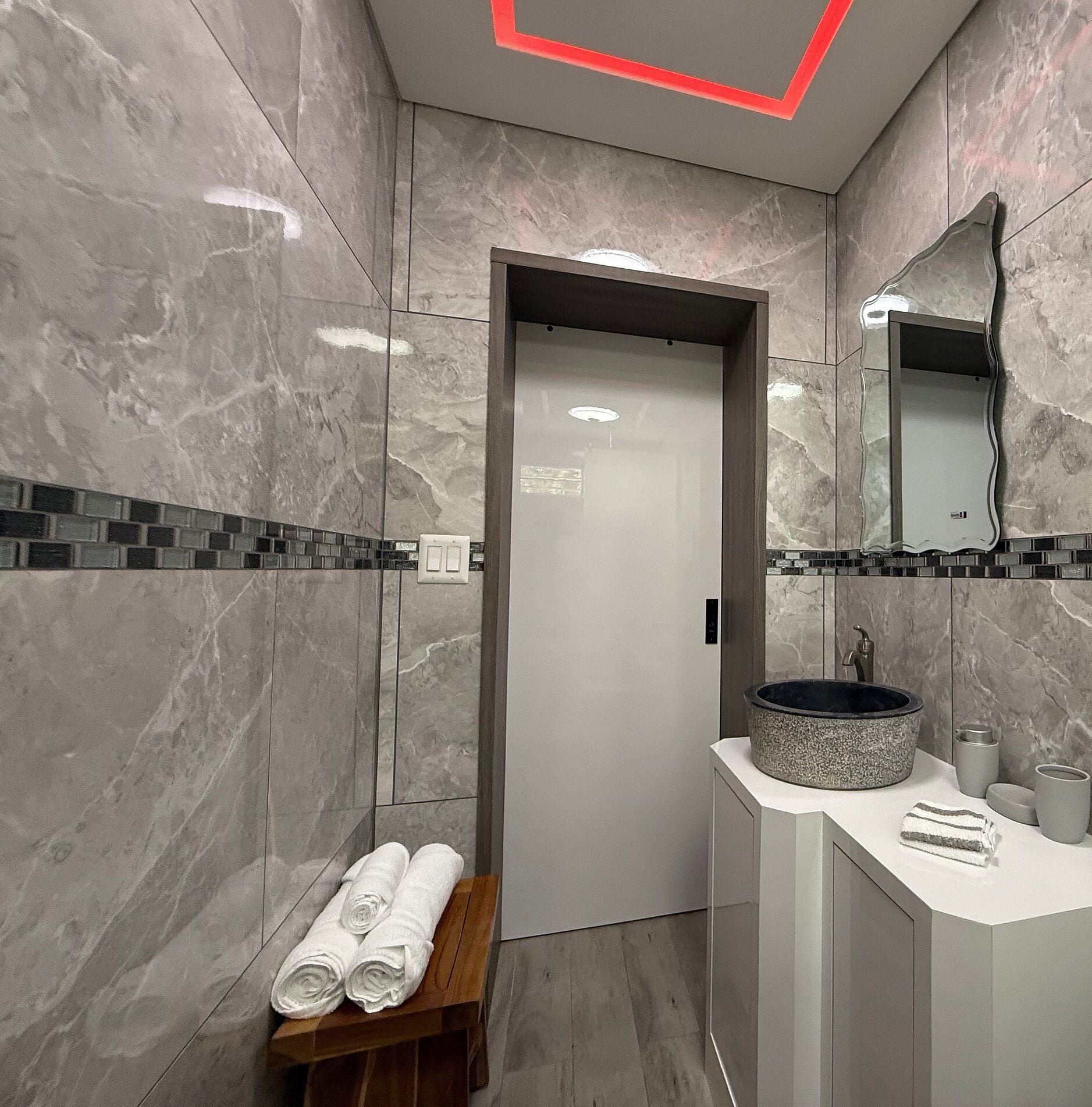 A modern bathroom with marble walls, a sink with a round basin, and a bench with towels.