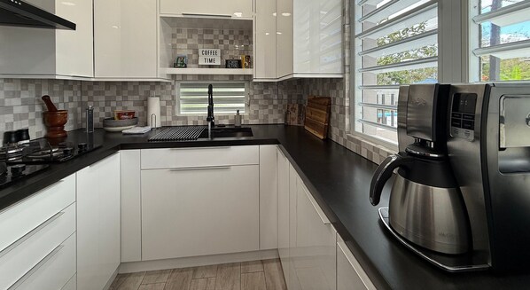 A modern kitchen with white cabinets, a black countertop, a coffee machine, and a window with a view of the outside. -  EcoMar Rentals Apartments (Cabo Rojo)
