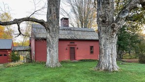 Una casa de madera roja con chimenea, rodeada de un césped cuidado y árboles grandes.