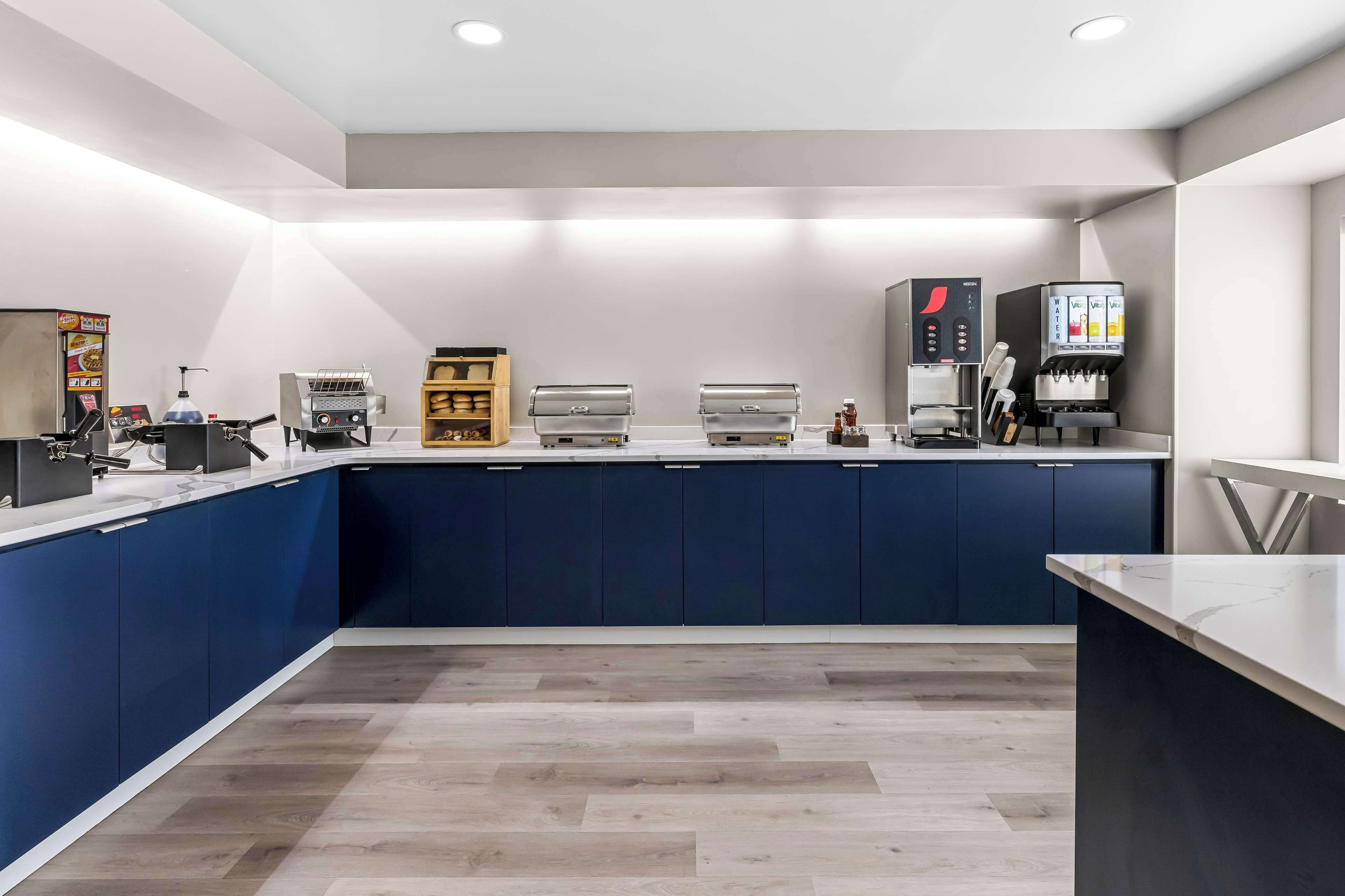 Een moderne keuken met blauwe kastjes, een marmeren aanrechtblad en diverse apparaten, waaronder een koffiezetapparaat en een warmhoudapparaat voor eten.