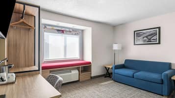 A room with a desk, a blue sofa, a window with a red bench, a framed picture on the wall, and a hanging coat rack.