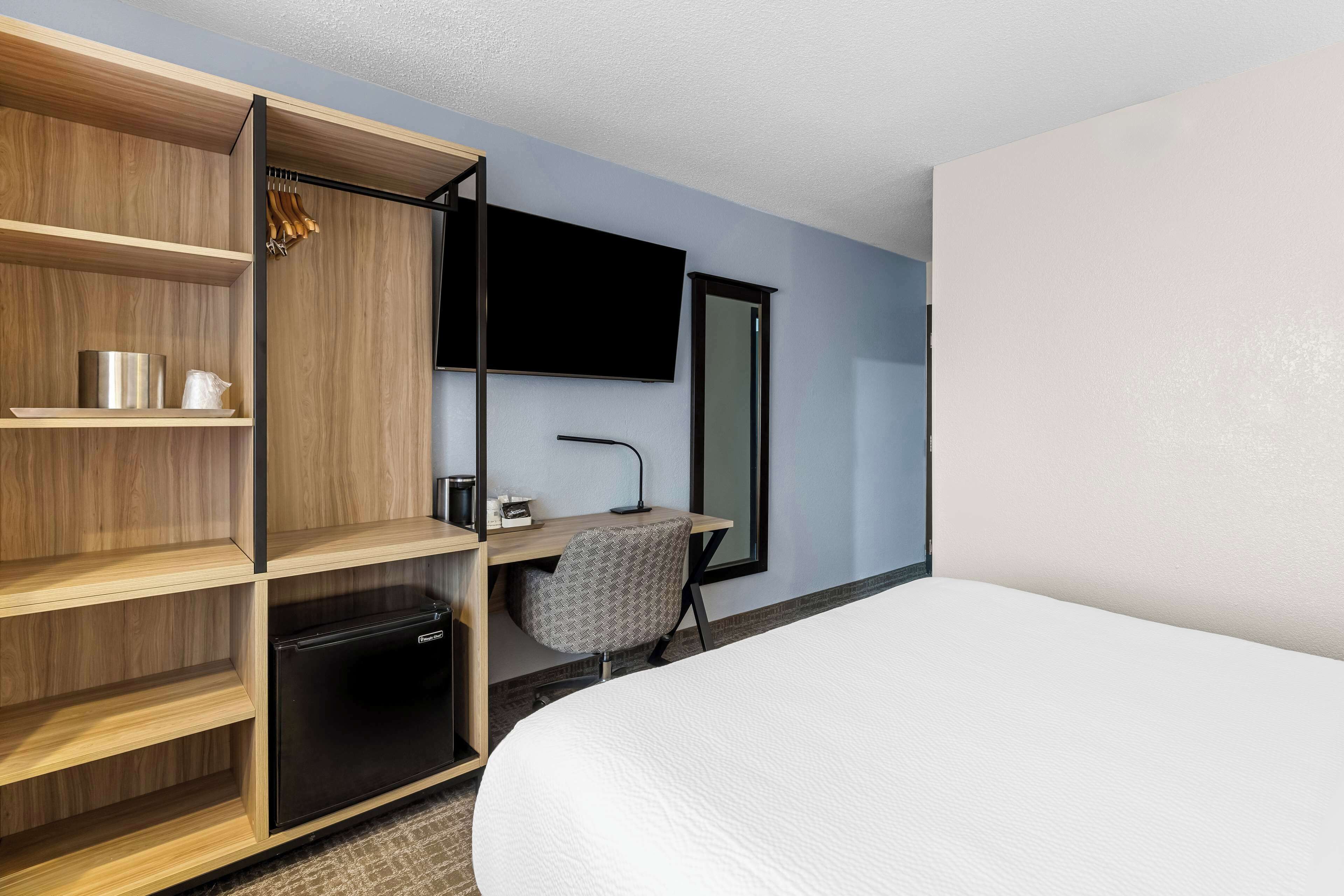 A hotel room with a bed, a desk with a chair, a TV, a wardrobe, and a small refrigerator.