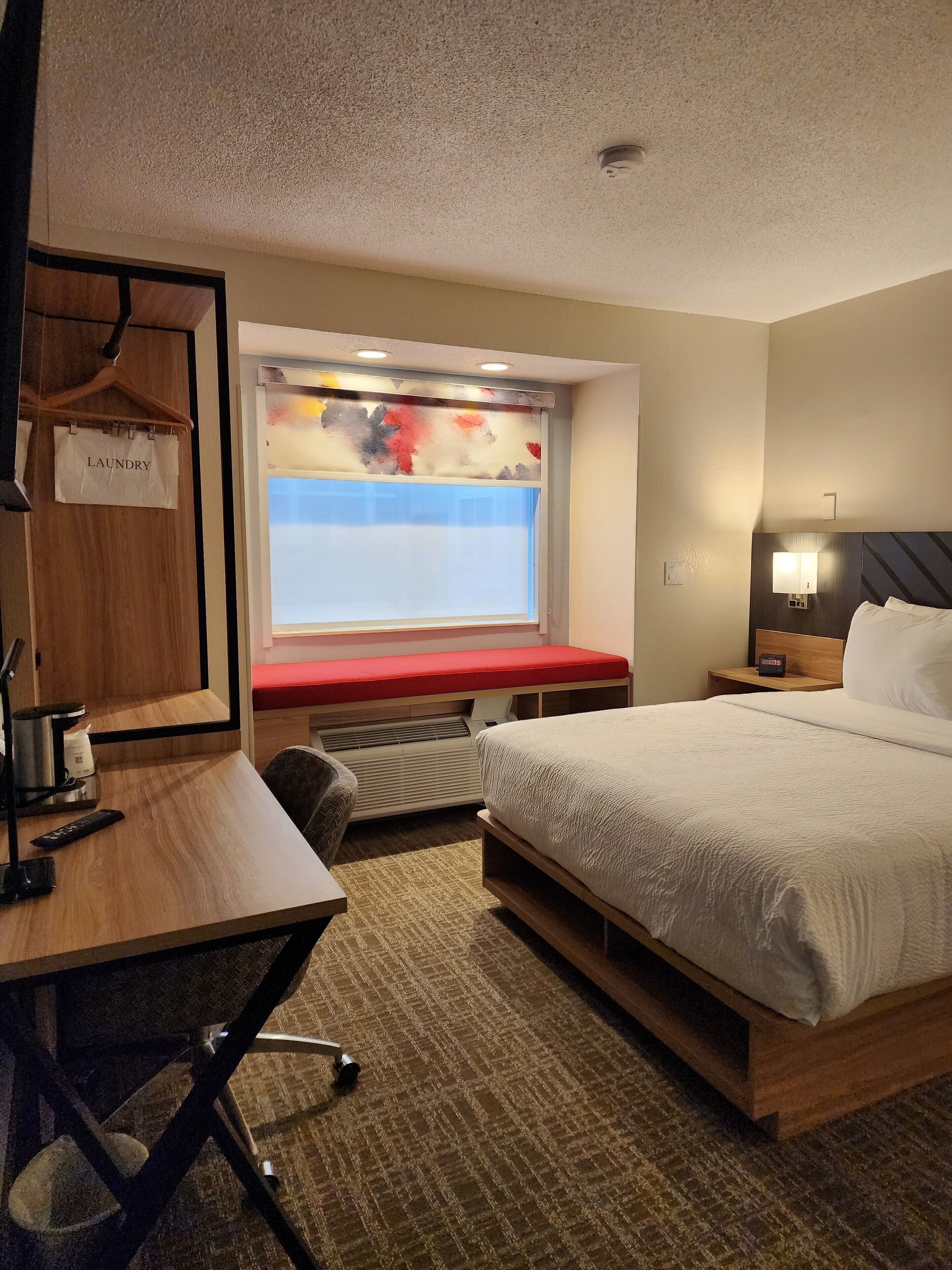 Une chambre d’hôtel comprenant un lit, un bureau avec une chaise, une fenêtre avec vue, une table de chevet avec une lampe et un panneau indiquant l’emplacement de la buanderie.