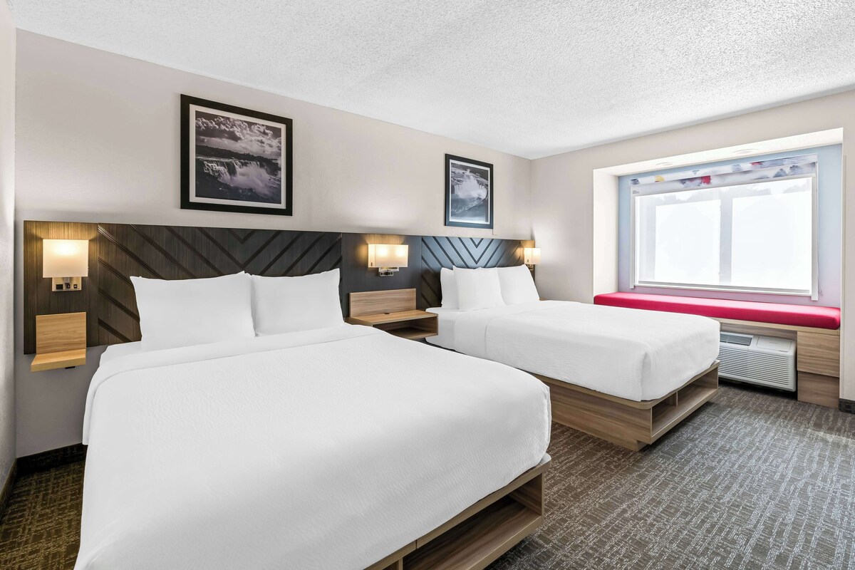 A hotel room with two beds, a large window with a view, and artwork on the walls.