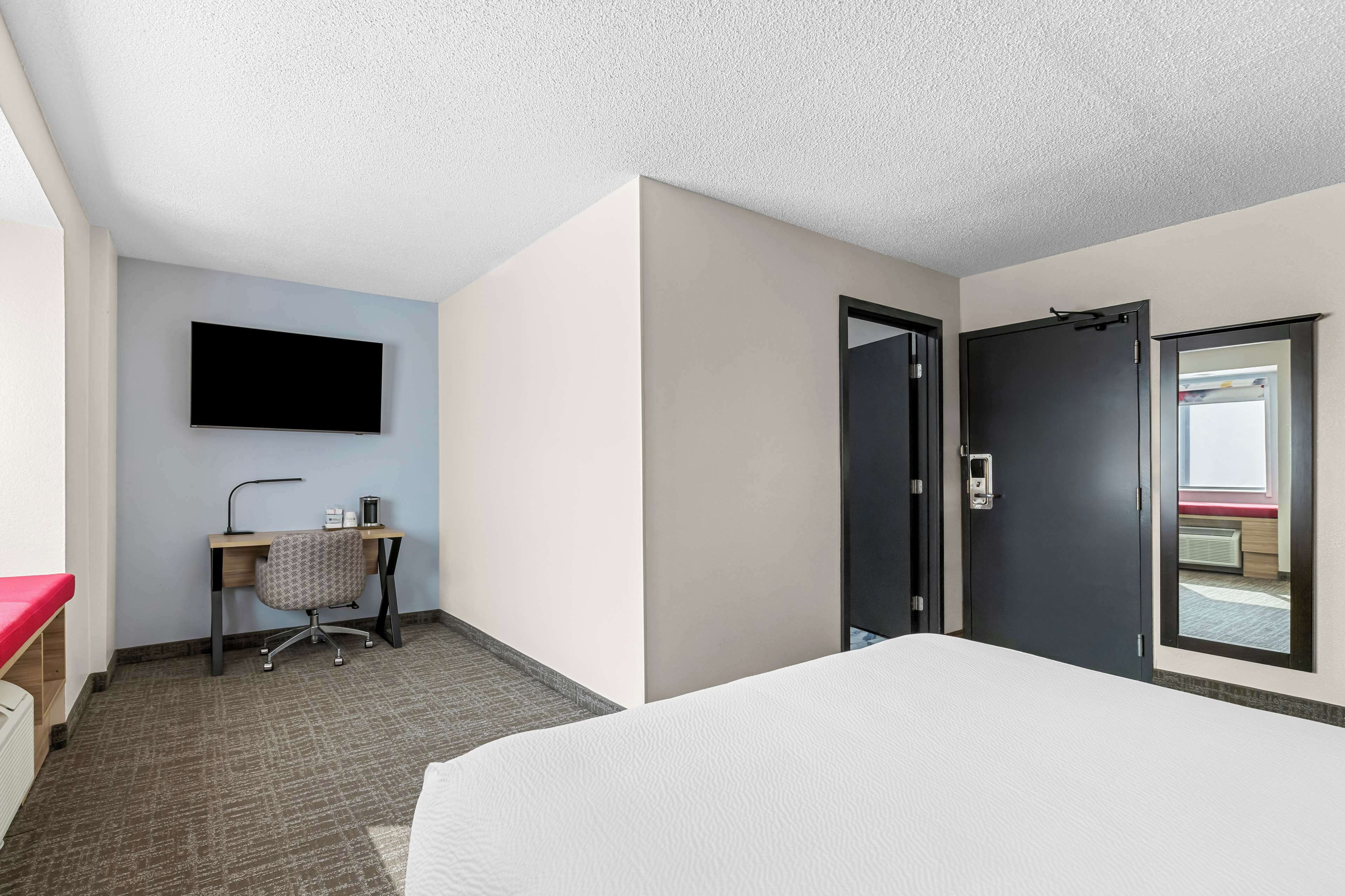 A hotel room with a bed, a desk with a chair, a flat-screen TV mounted on the wall, and a door leading to a bathroom.