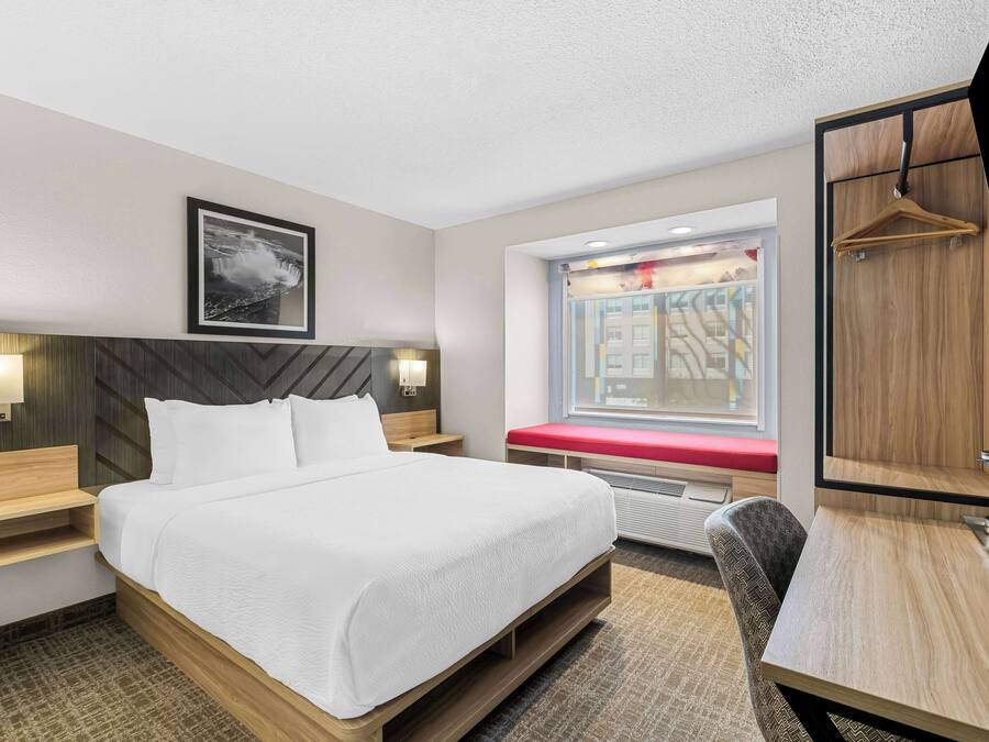 A hotel room with a large bed, bedside tables, a desk, a chair, a window with a view, and a mounted TV.